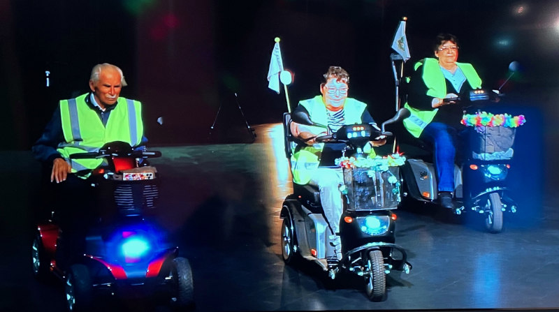3 selbstfahrende Rollstühle – Sccoter – mit ihren Fahrern nebeneinander auf einer Bühne.